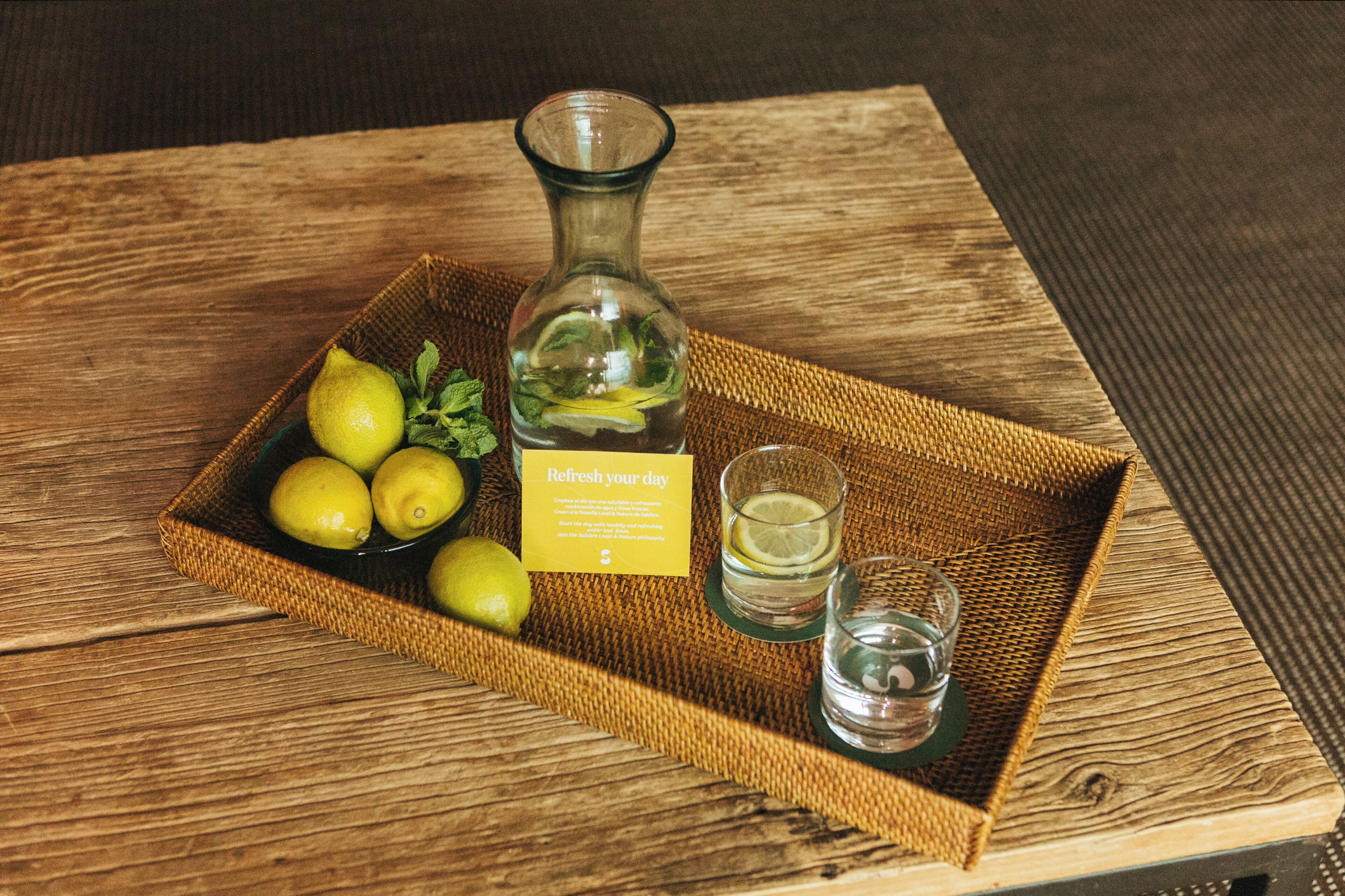 A wicker tray with lemons, a bottle, and two shot glasses on a wooden table.