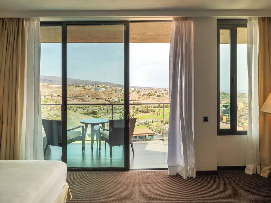 Habitación de hotel con un amplio ventanal corredero que da a un balcón con mesa y sillas, con vistas al paisaje.
