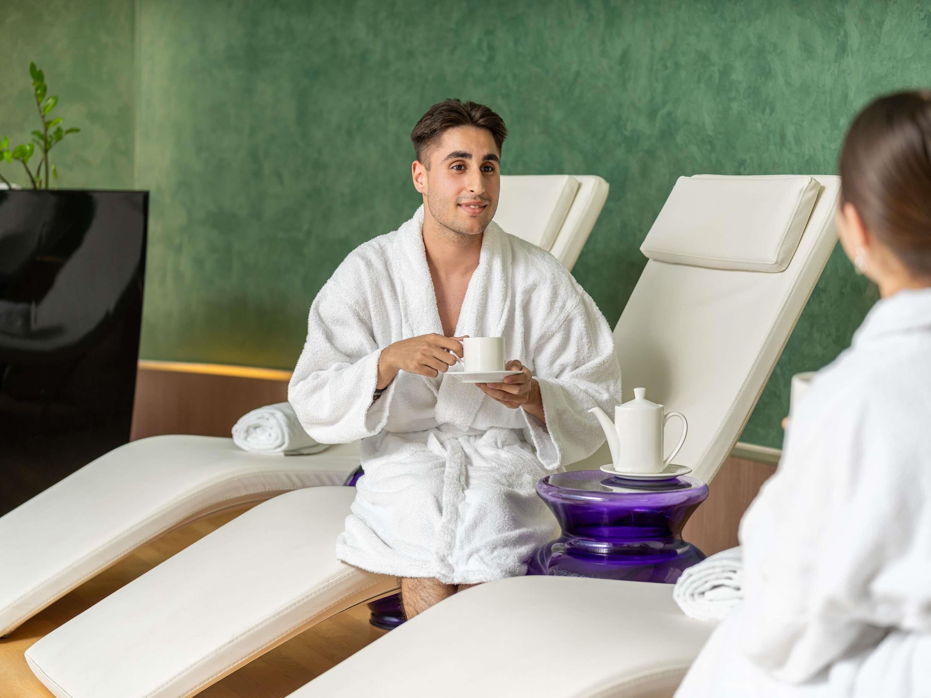 Un entorno de spa con una persona vestida con una bata blanca sosteniendo una taza, una silla de masaje y una camilla de masajes morada.