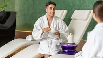 Un entorno de spa con una persona vestida con una bata blanca sosteniendo una taza, una silla de masaje y una camilla de masajes morada.