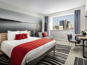 A hotel room with a bed, a desk, a chair, a view of a cityscape, and a painting on the wall. - Novotel Bucharest City Centre (Bucharest)