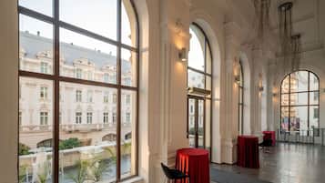 A spacious hotel lobby with large arched windows, a central red table, and a view of a historic building.