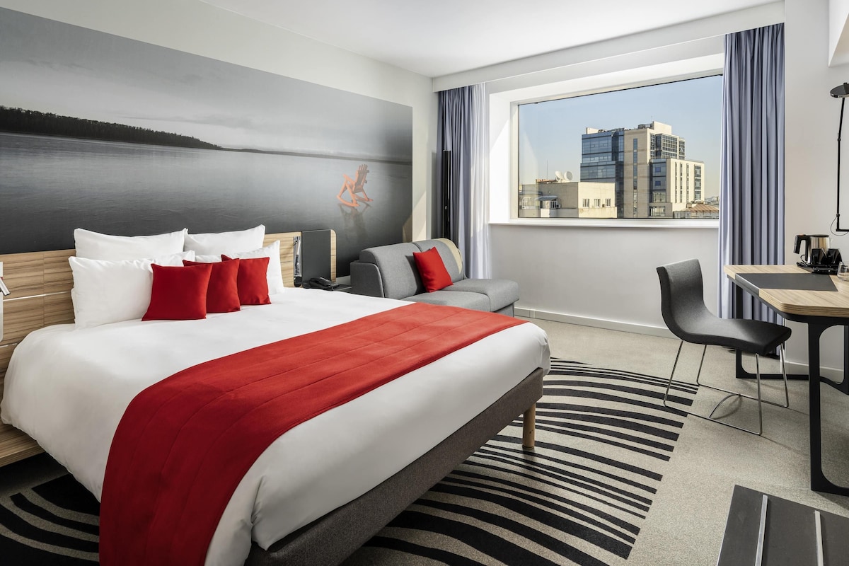 A hotel room with a bed, a desk, a chair, a view of a cityscape, and a painting on the wall.