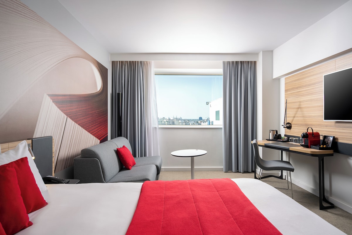 A hotel room with a bed, a desk, a chair, a small table, and a TV.
