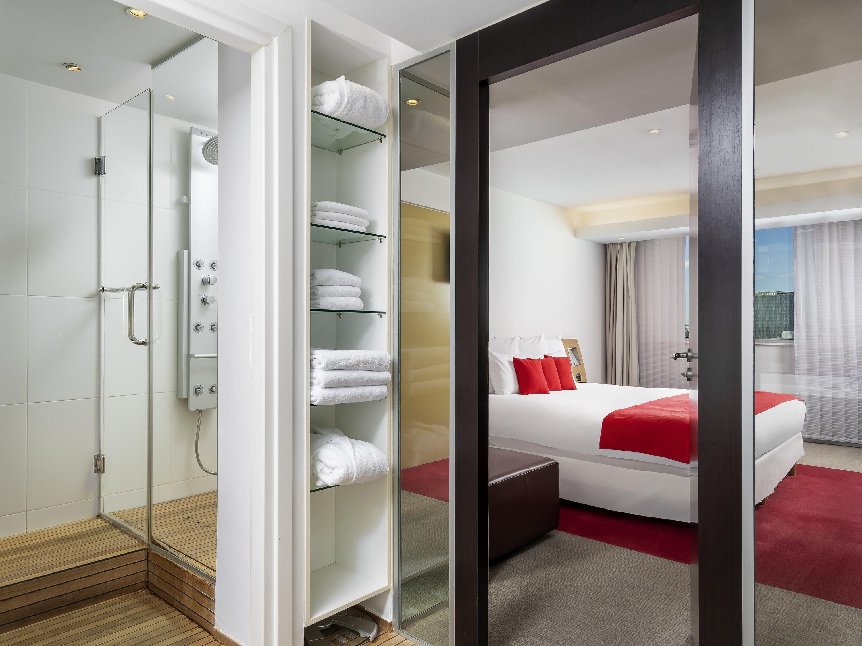 Une chambre d’hôtel dotée d’une douche à parois de verre, d’une coiffeuse avec un plateau en verre, d’un lit avec une literie rouge et offrant une vue sur la ville.