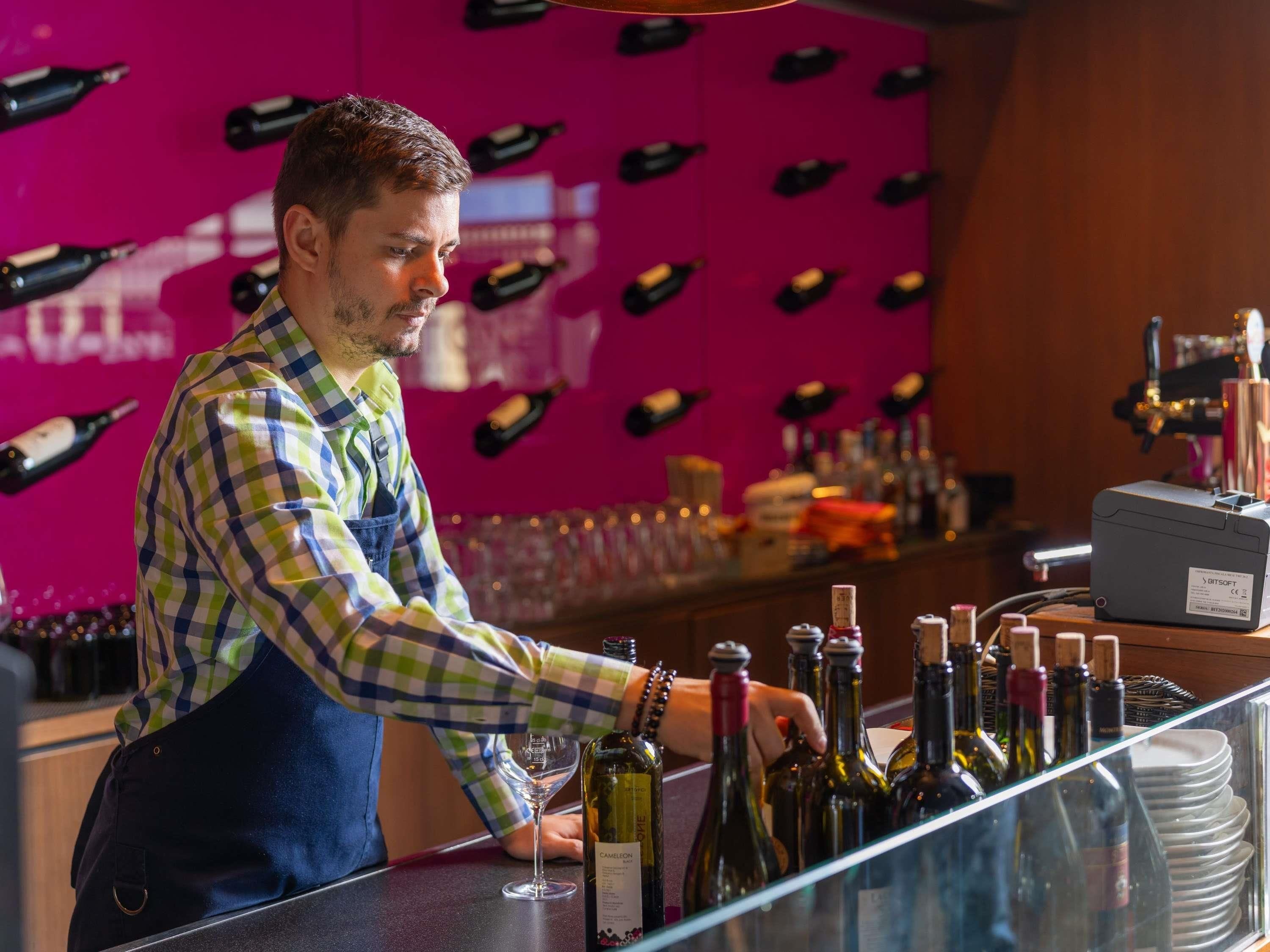 A bar area with a bartender preparing drinks. There are multiple bottles of wine and a wine glass on the counter.