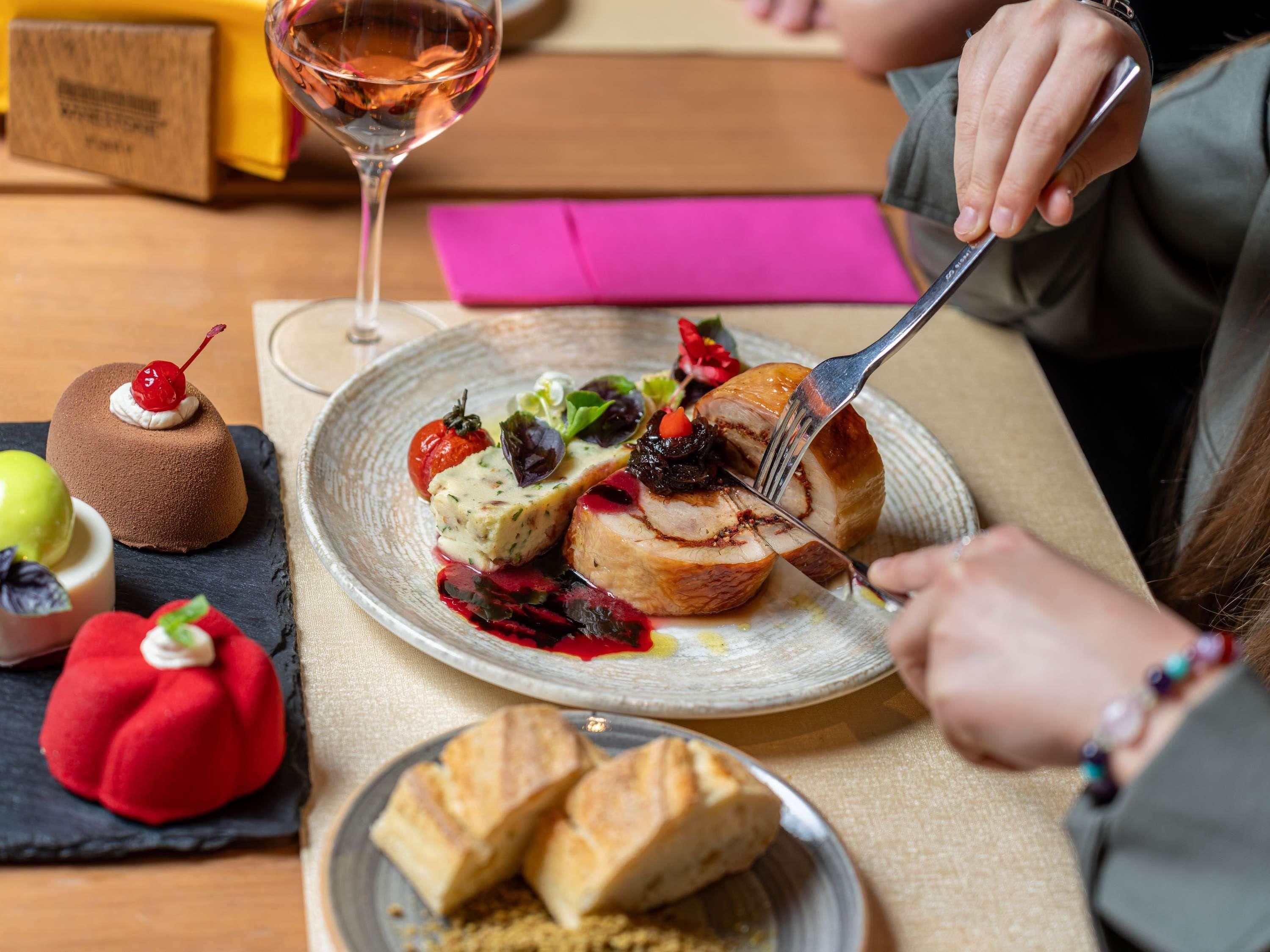 A dining table with a plate of food, a glass of wine, and a person cutting meat.