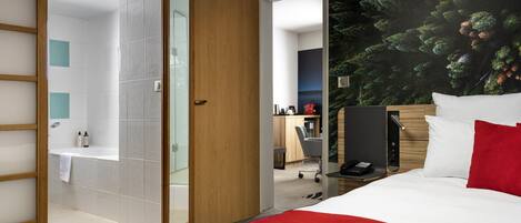 A hotel room with a bed, a shower area, a desk, and a large window with a forest view.