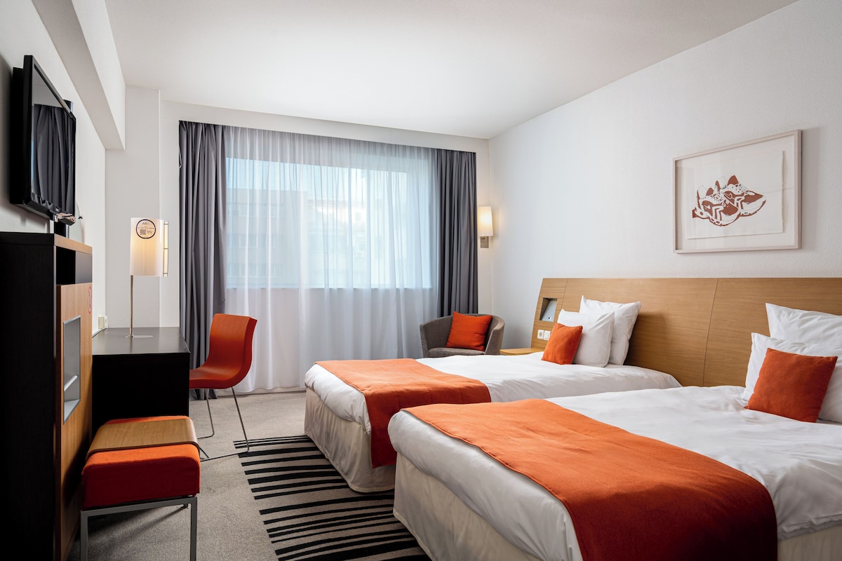 A hotel room with two beds, a desk, a chair, a TV, and a large window with curtains.