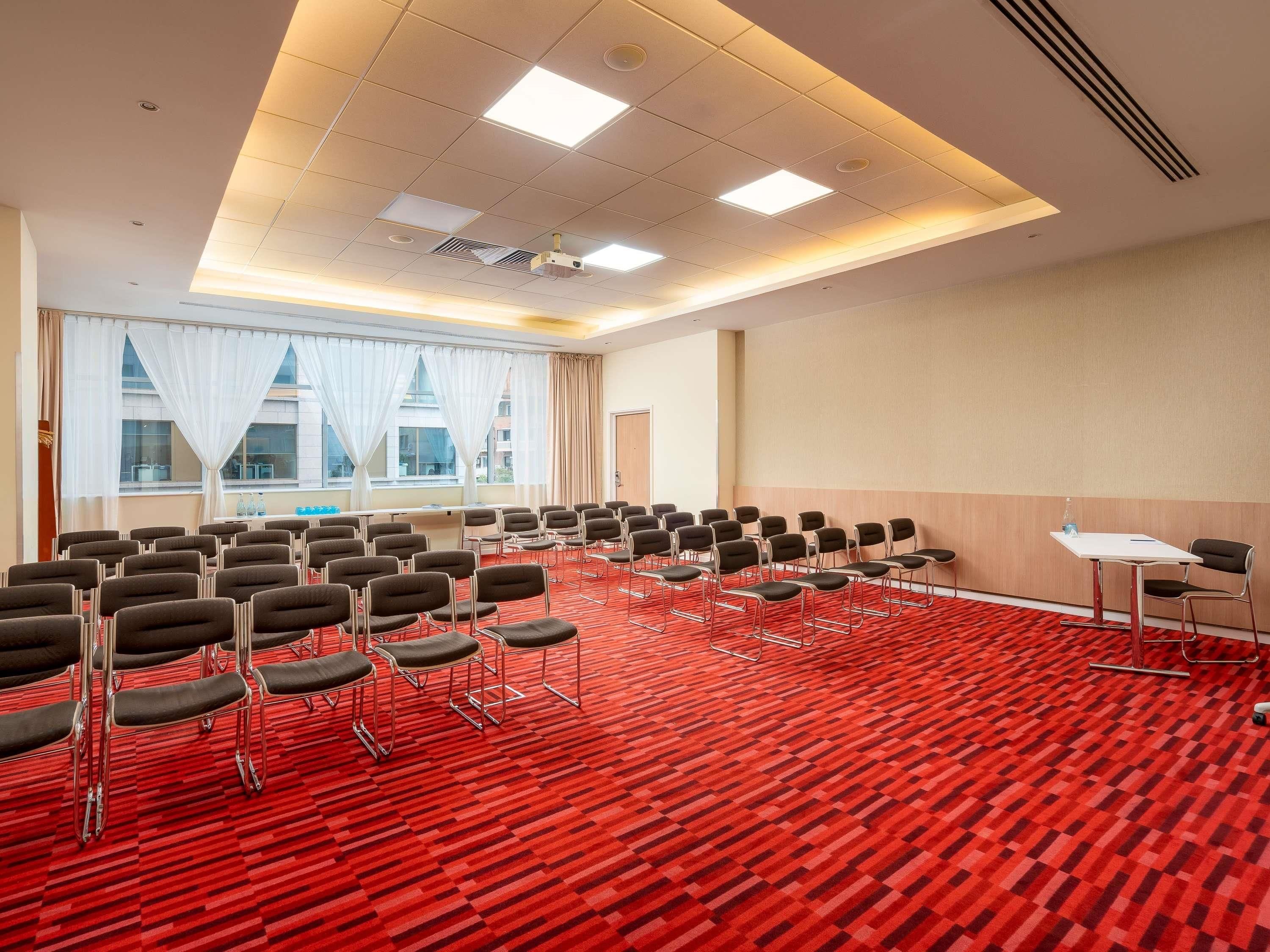 Un salón de conferencias con filas de sillas, un proyector y un amplio ventanal con cortinas.