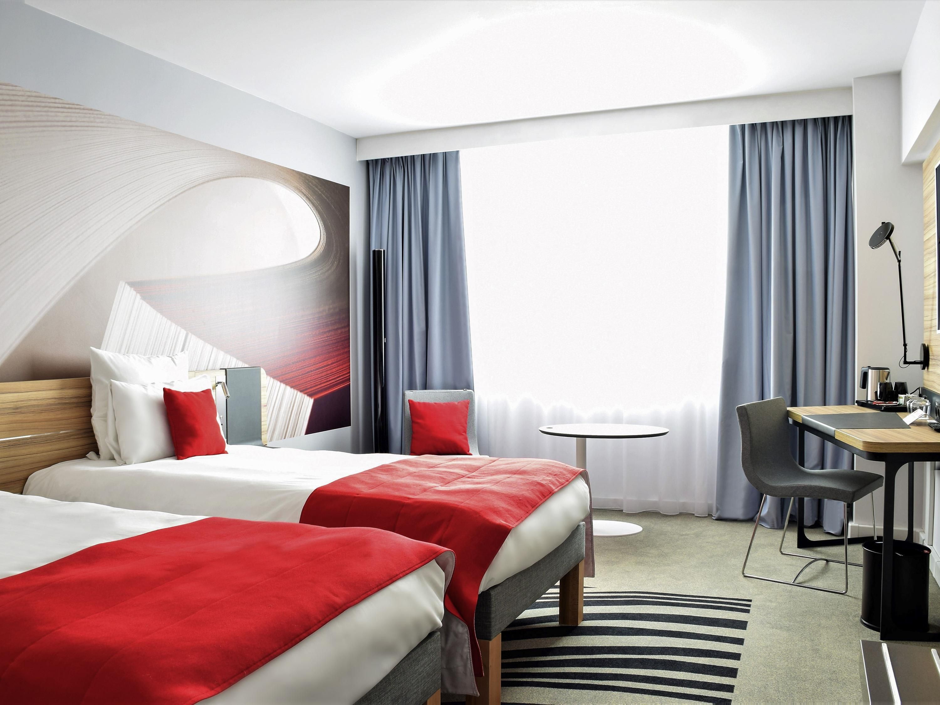A hotel room with two beds, a desk, a chair, a small round table, and a large window with curtains.