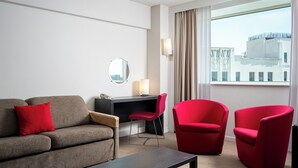 Habitación de hotel con un sofá, dos sillas rojas, un escritorio y un ventanal con vistas a edificios.