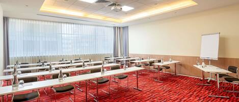 Un salón de conferencias con alfombra roja, varias mesas con sillas, una pizarra y un amplio ventanal con cortinas.