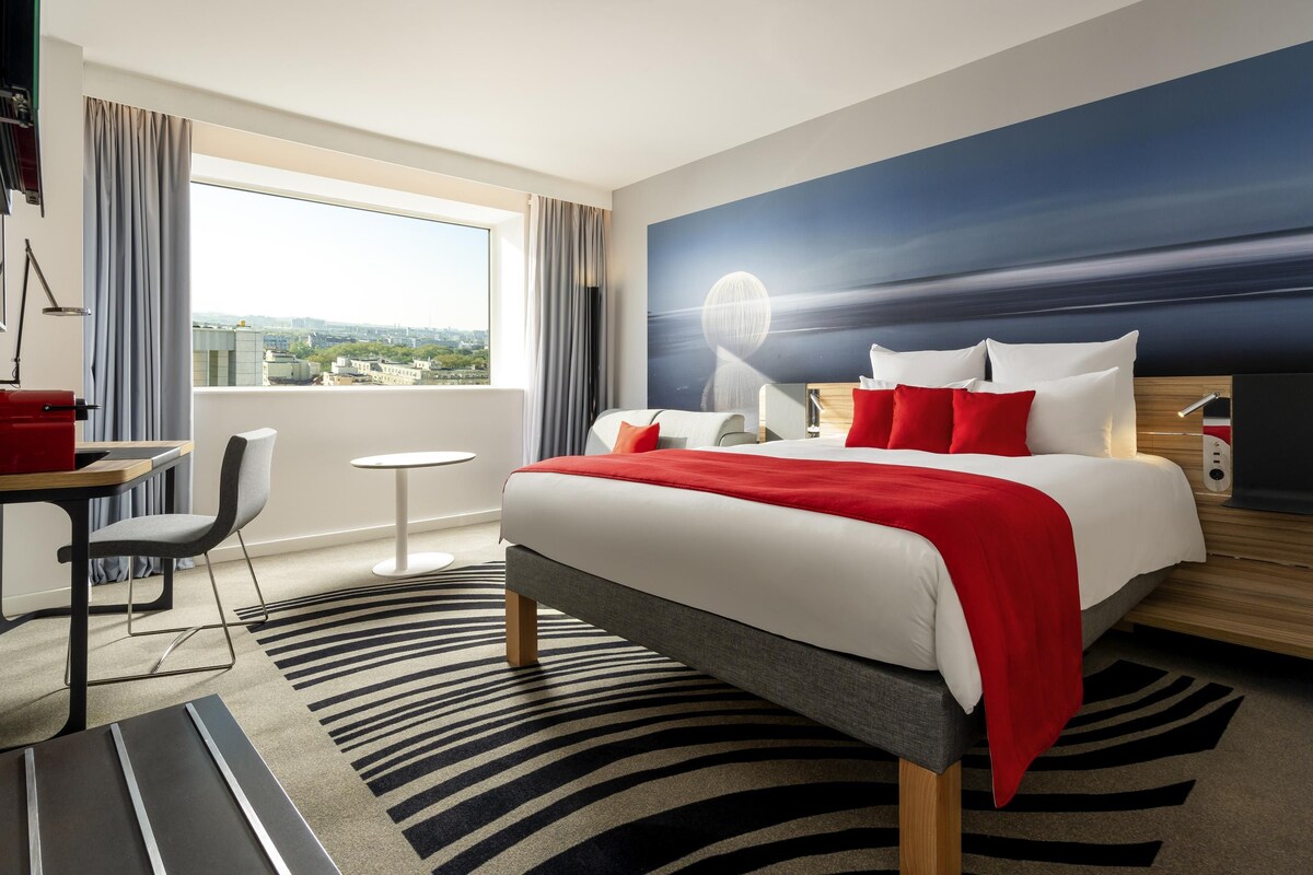 A hotel room with a bed, a desk, a chair, a small table, and a view of the city.