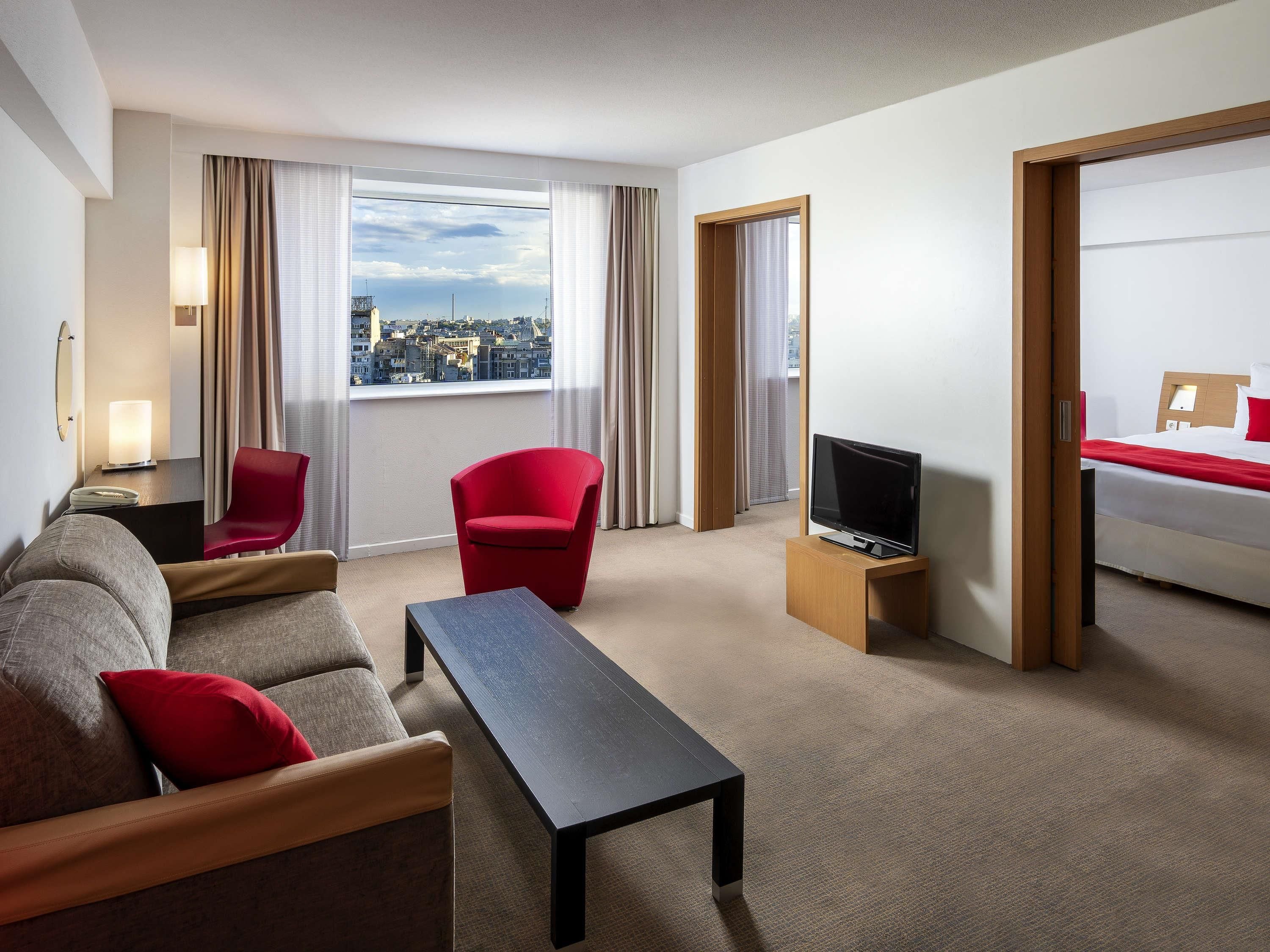 A hotel room with a sofa, a red chair, a TV on a wooden stand, and a bed with a red pillow.