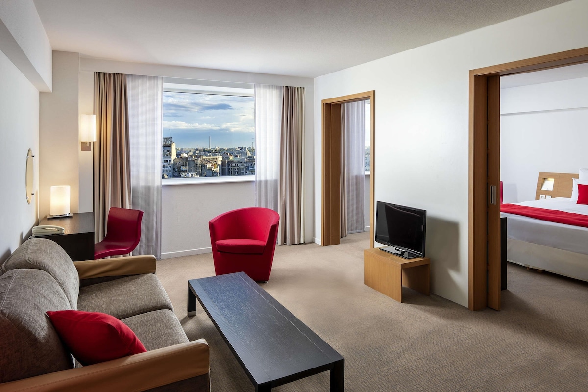 A hotel room with a sofa, a red chair, a TV on a wooden stand, and a bed with a red pillow.