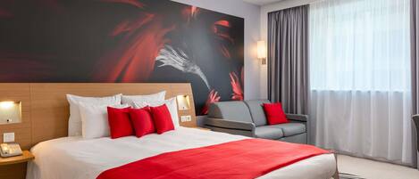 Habitación de hotel con una cama grande, un sofá gris, una manta roja y un ventanal con cortinas.