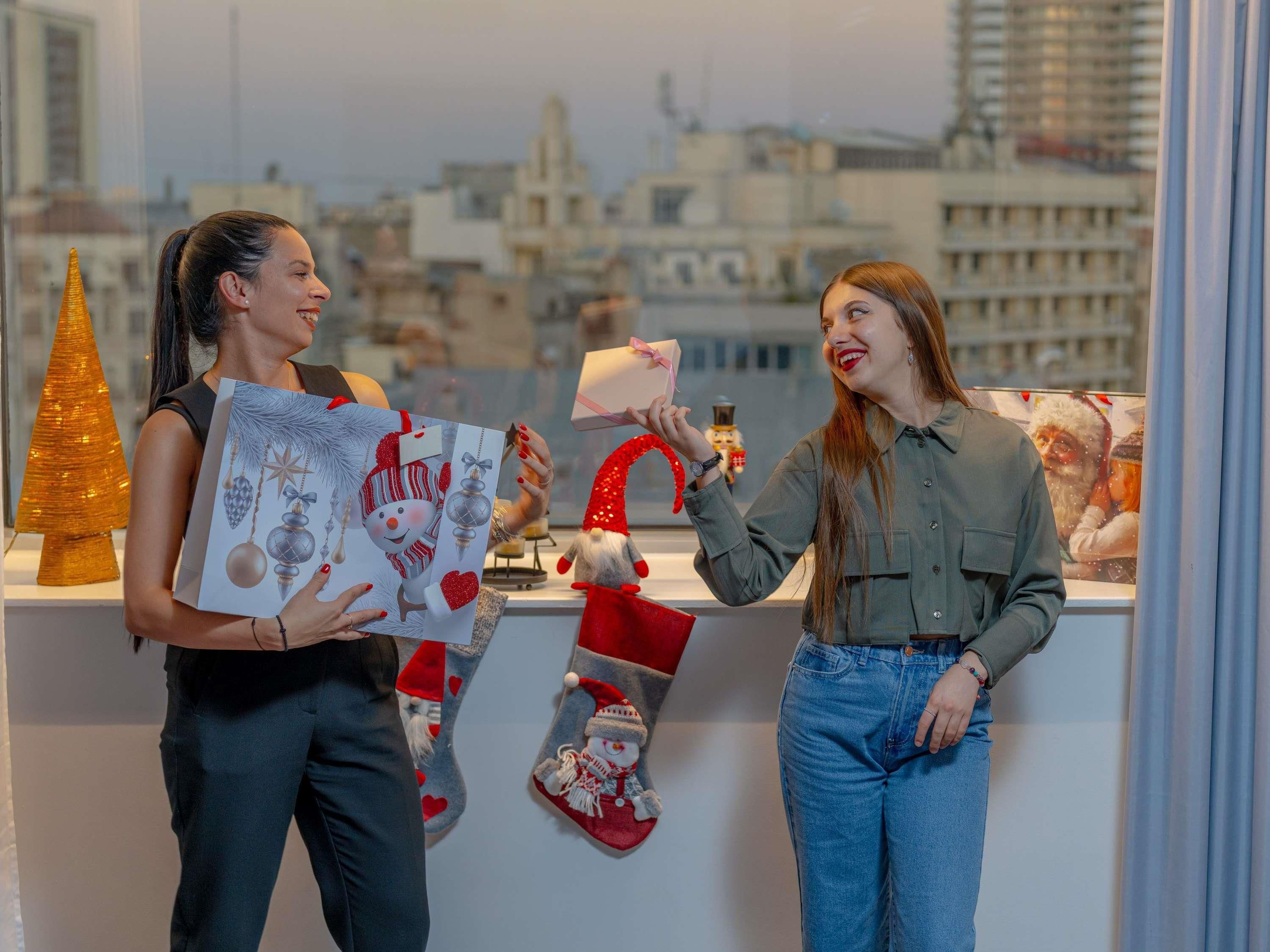 Two people exchanging gifts at a counter with a city view, a Christmas tree, and festive decorations.