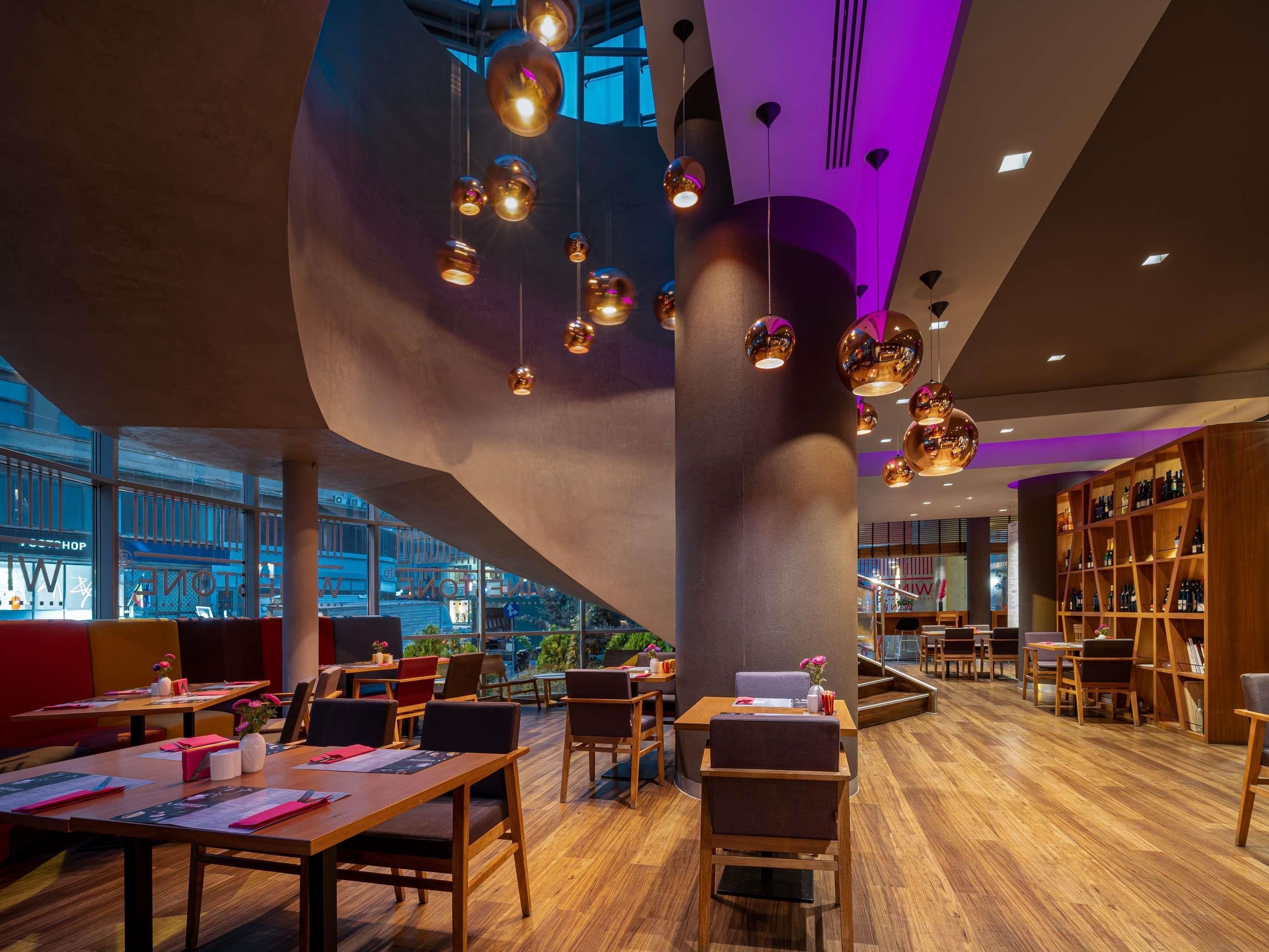 Un interior moderno con mesas y sillas de madera, una estantería grande e iluminación colgante.