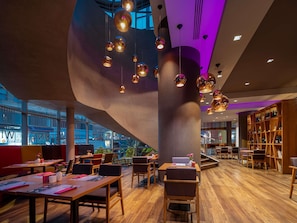 Un interior moderno con mesas y sillas de madera, una estantería grande e iluminación colgante.