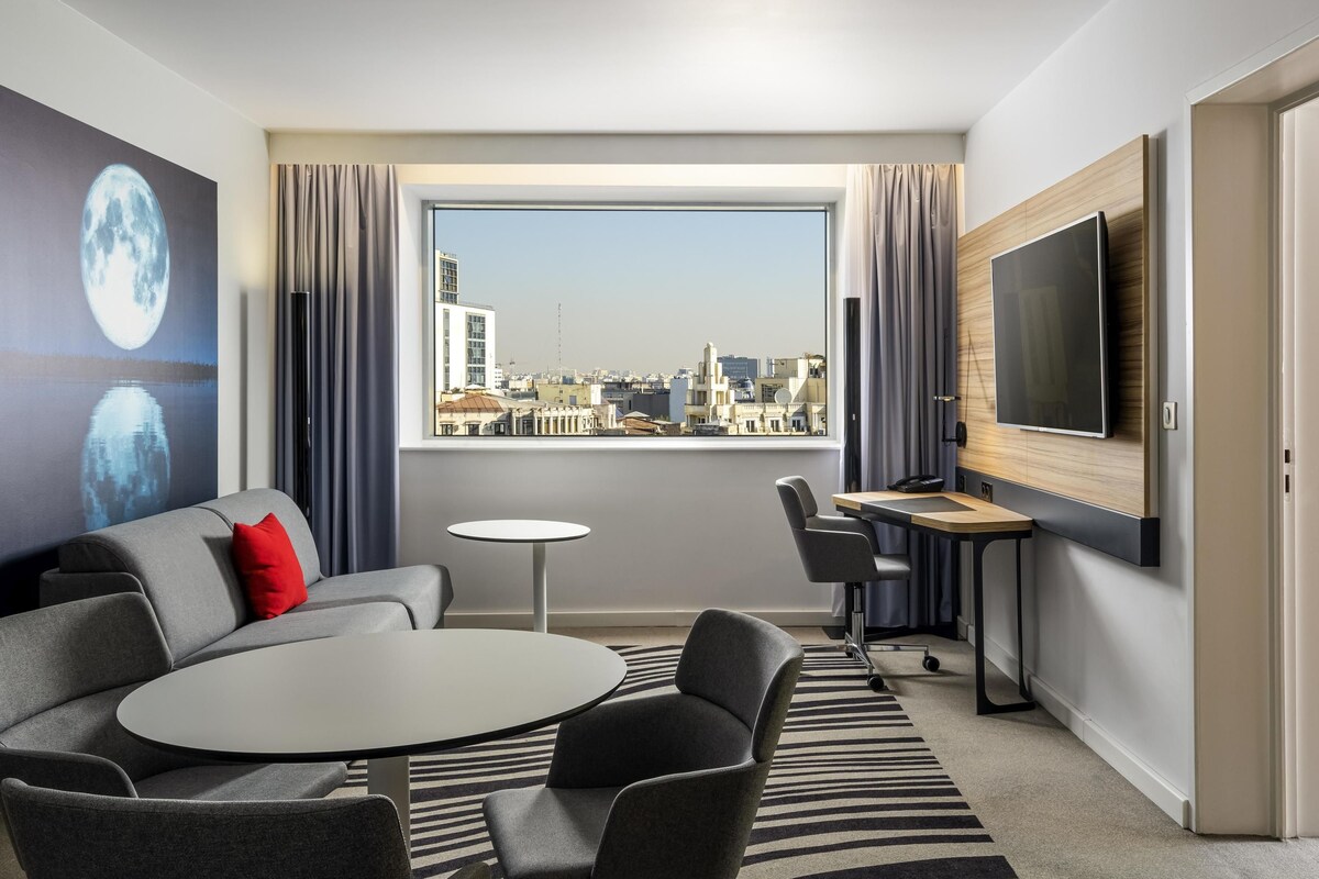 A modern hotel room with a city view, a sofa, a round table, and a TV.