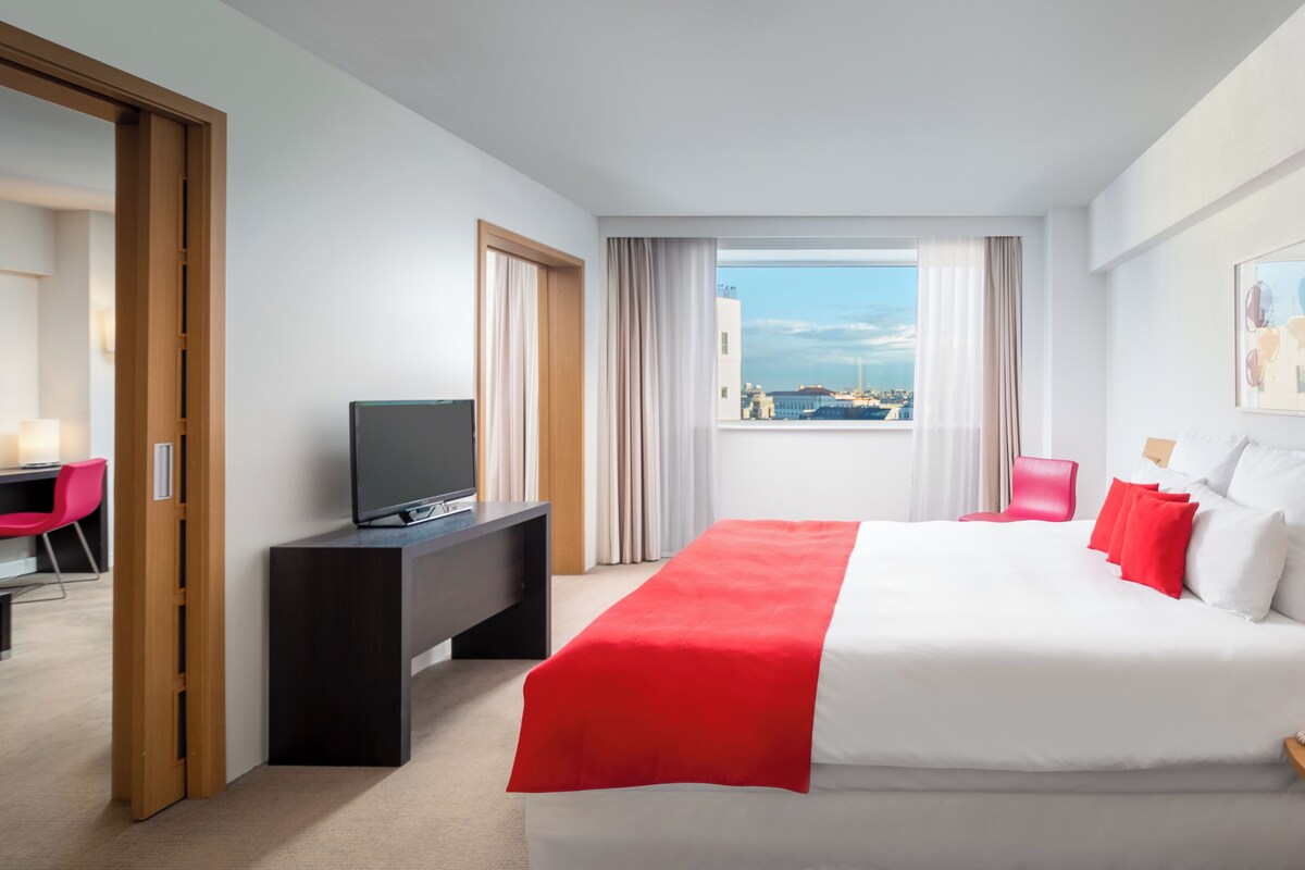 A hotel room with a bed, a TV on a desk, a view of the city, and a red chair.