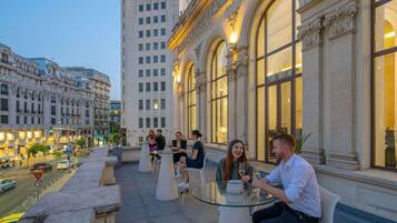 A rooftop terrace with outdoor seating, a view of a historic building, and a busy street below.