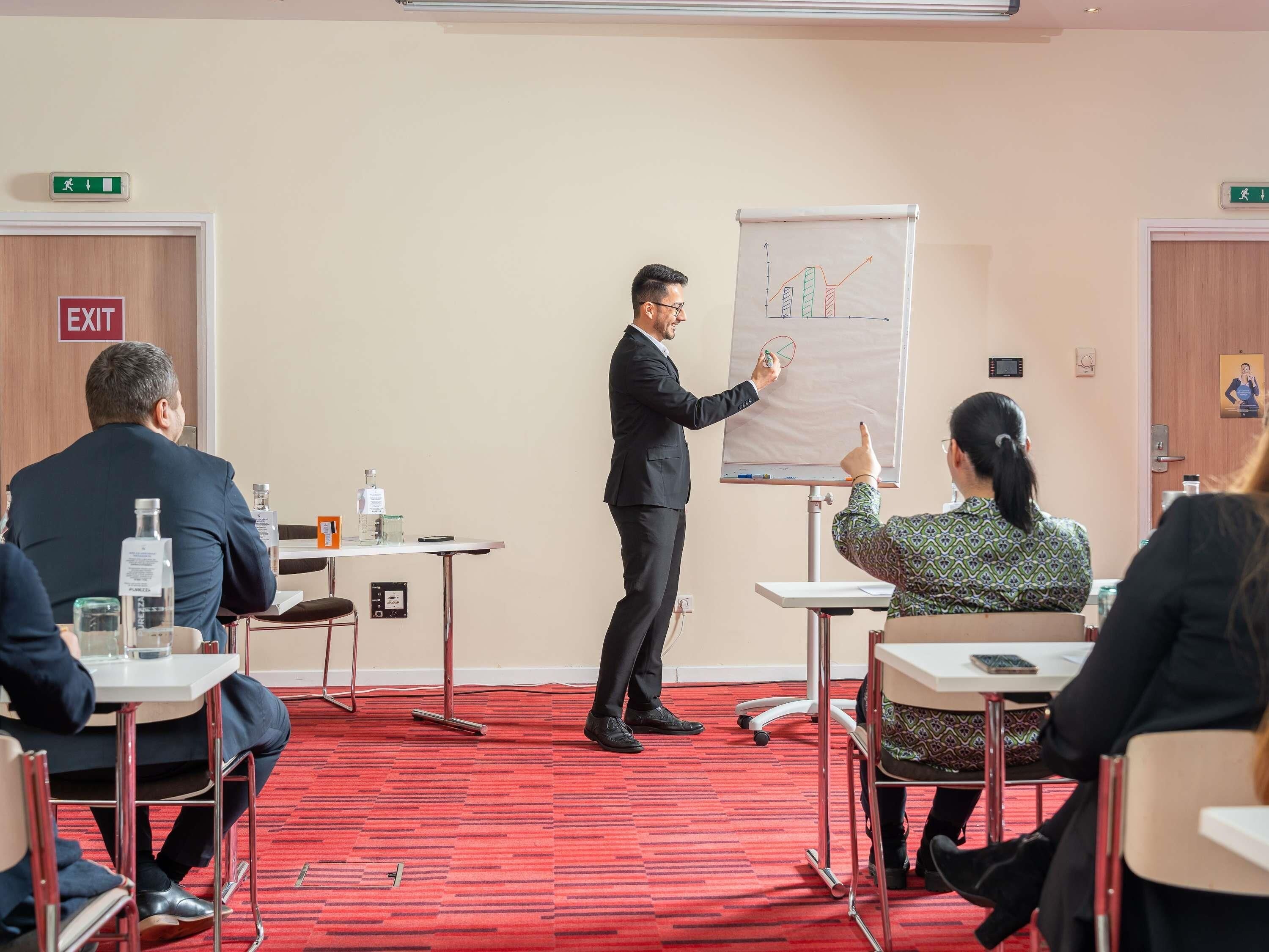A conference room with a presenter, attendees seated at tables, and a whiteboard with a chart.