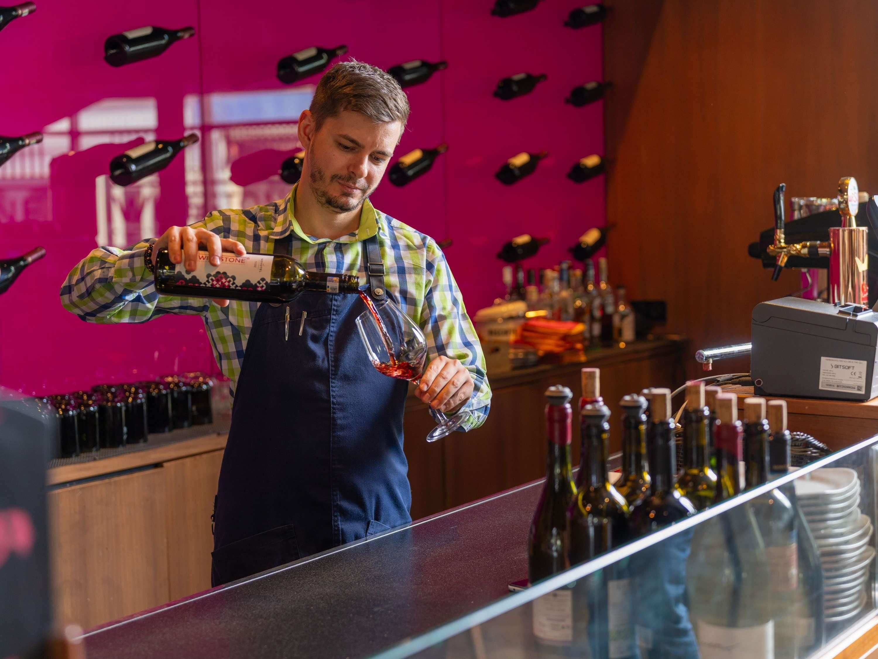 A person pouring wine into a glass at a bar with a pink wall and wine bottles in the background.
