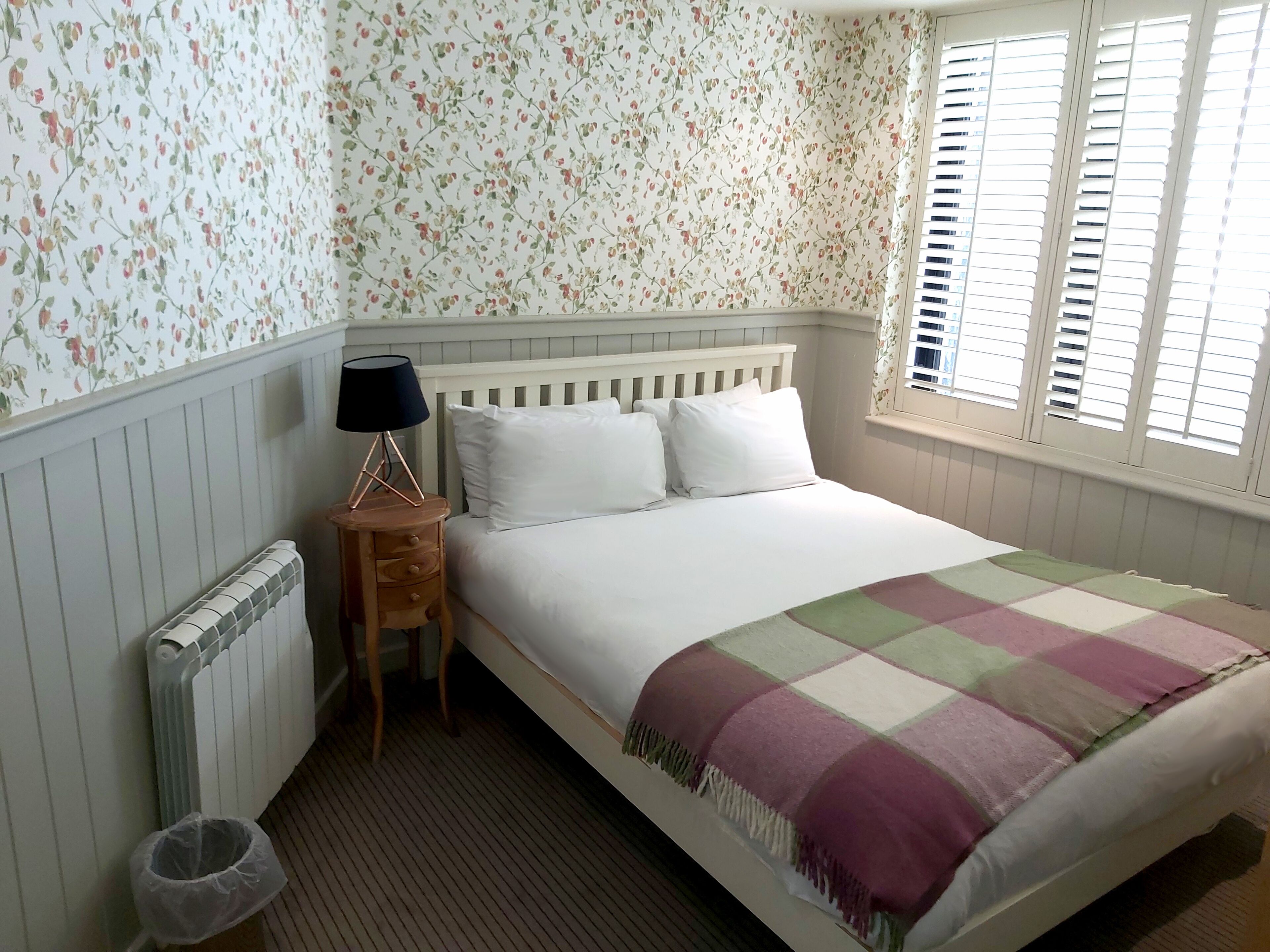 A bedroom with a bed, a nightstand, a lamp, a radiator, a window with shutters, and a trash bin.