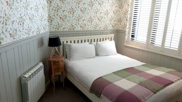 A bedroom with a bed, a nightstand, a lamp, a radiator, a window with shutters, and a trash bin.