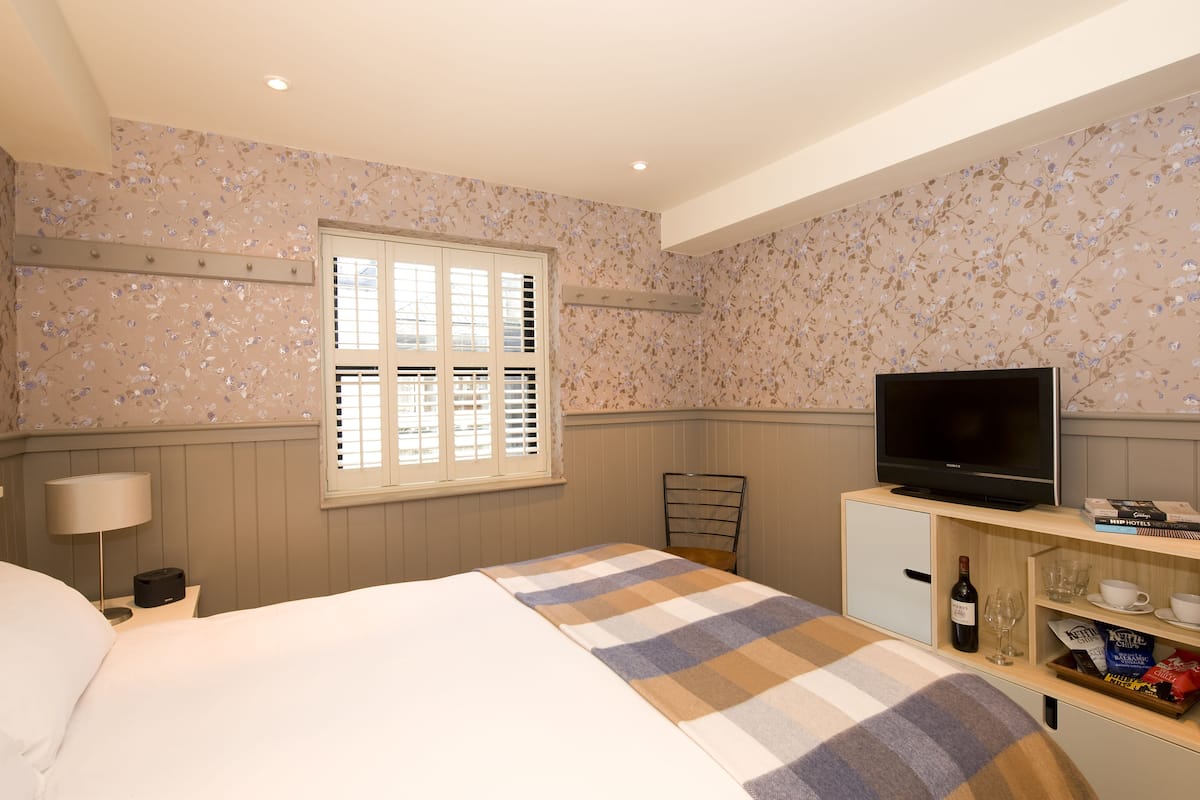 Ein Hotelzimmer mit Bett, Fernseher, einem kleinen Schrank, einem Fenster mit Rollläden und einer Wand mit Blumenmuster.