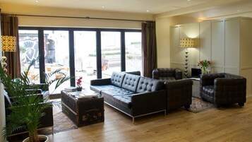 A spacious living room with a large sofa, a smaller armchair, and a coffee table. There is a potted plant and a lamp on a side table.