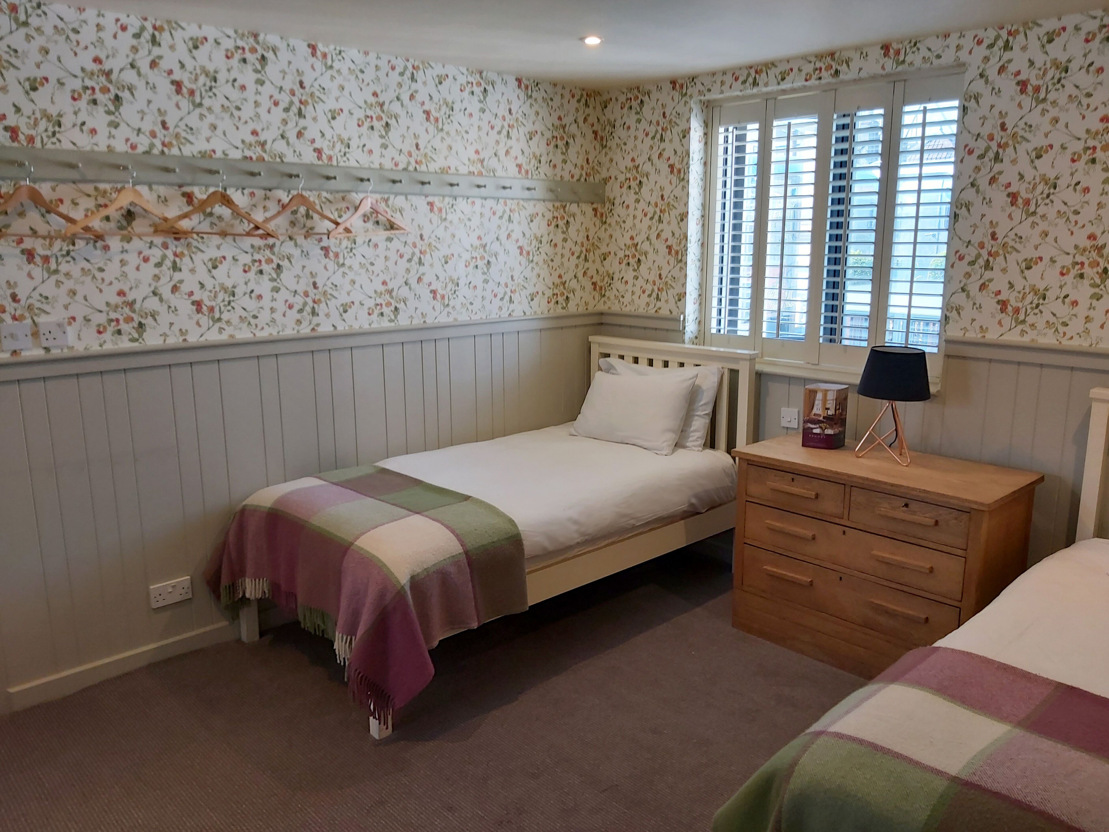A bedroom with a single bed, a wooden dresser, a nightstand, a lamp, and a window with shutters.