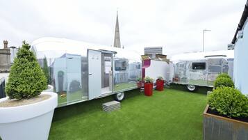 A row of trailers with a grassy area in between, a church spire in the background, and potted plants around.