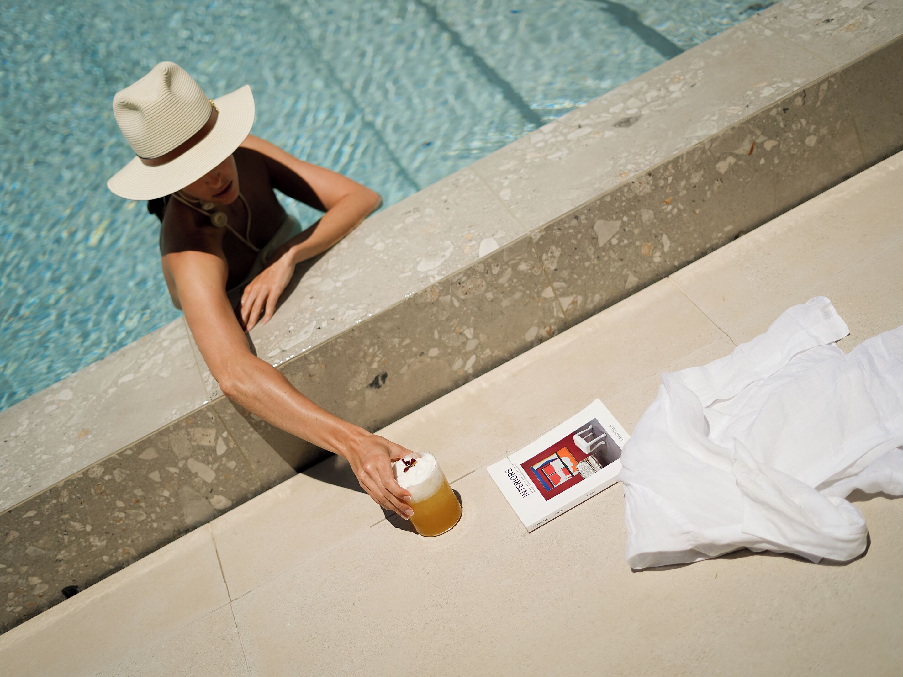 A person by a poolside with a drink and a magazine.