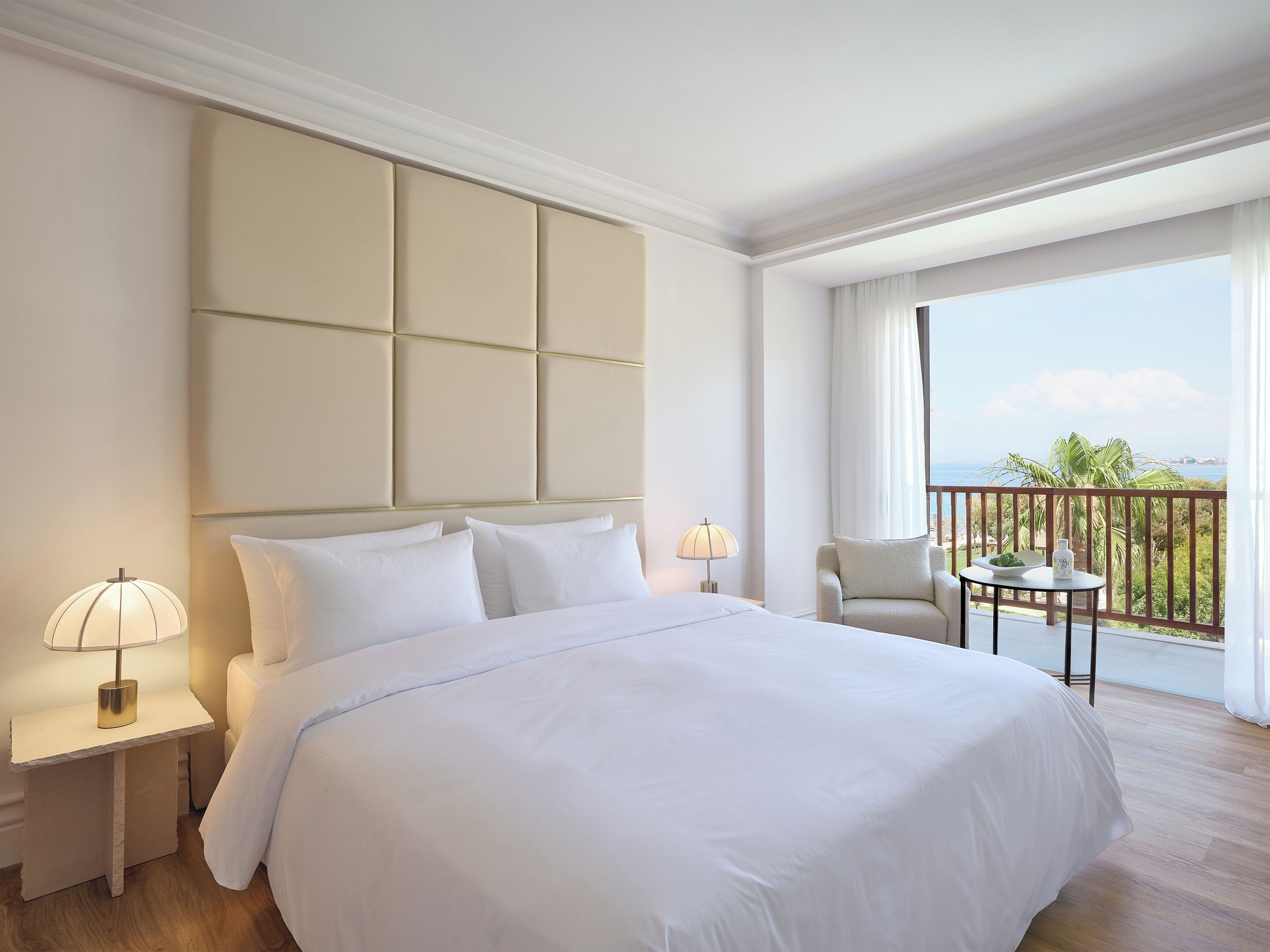 Habitación de hotel con una cama grande, una mesita de noche con lámpara, una silla y un balcón con vista al océano.