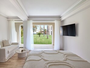 A modern hotel room with a sofa, a round coffee table, a flat-screen TV, and a view of a well-maintained garden. - Grecotel Filoxenia (Kalamata)