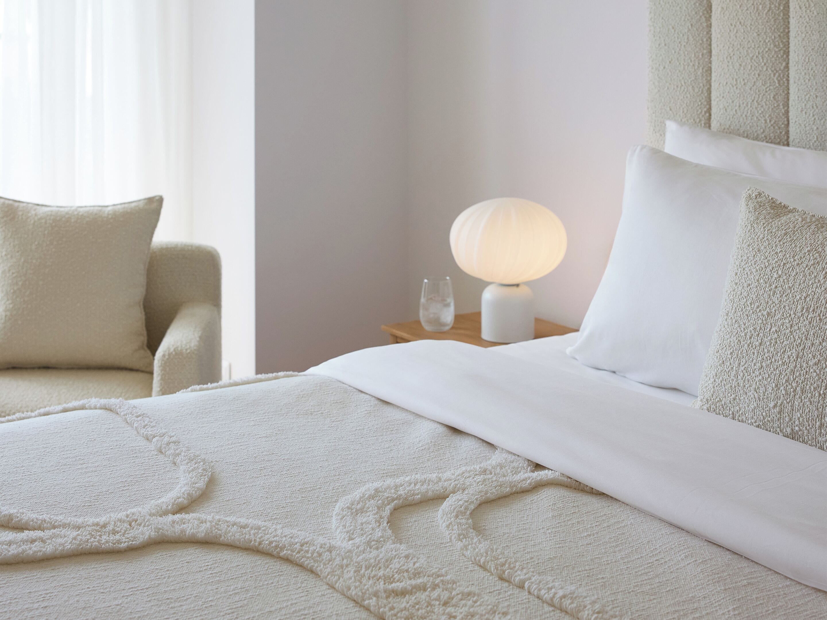 A neatly made bed with a fluffy white blanket and pillows, a bedside lamp, and a glass of water on a wooden nightstand.