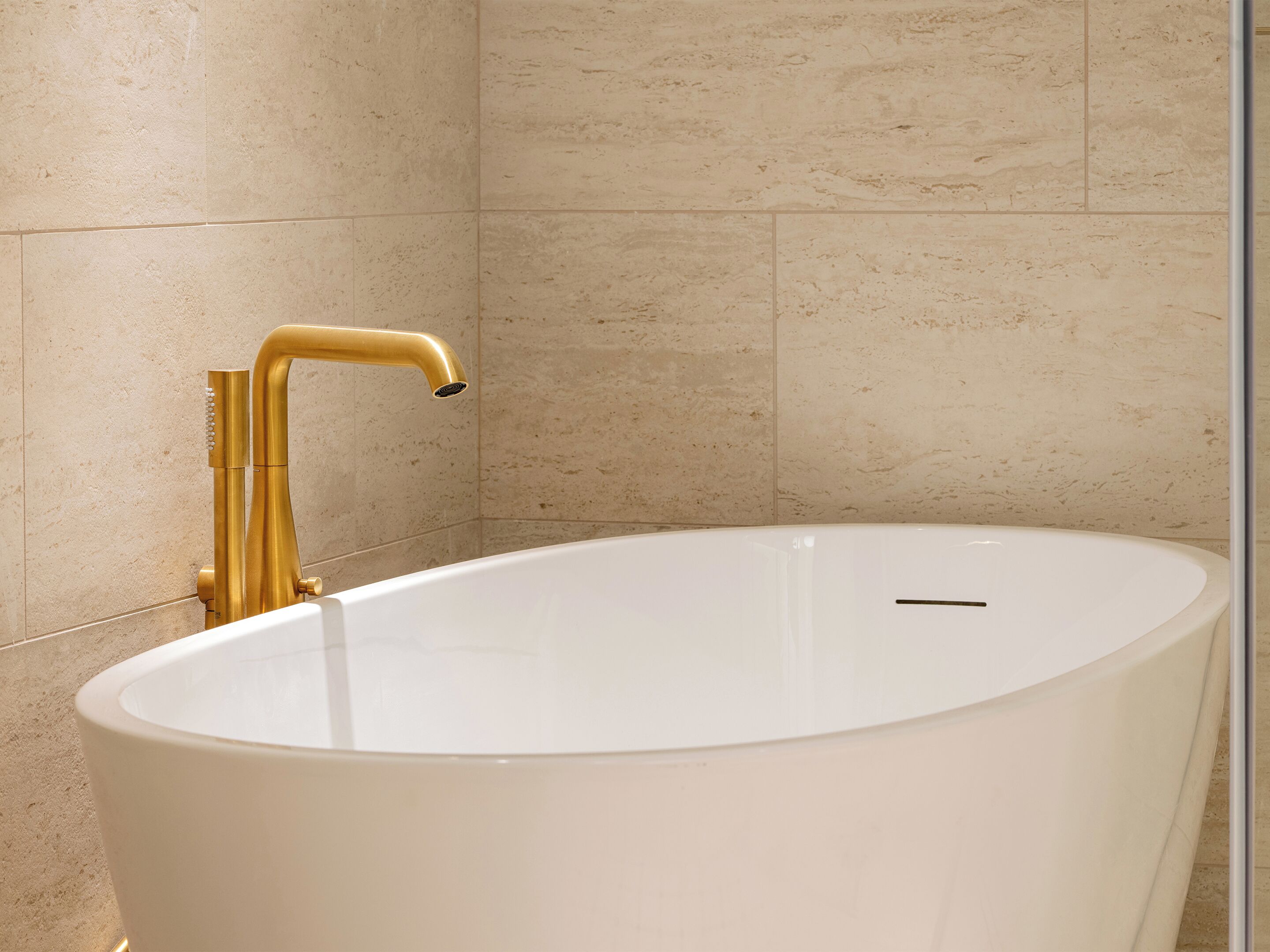 A modern bathroom with a freestanding bathtub, gold fixtures, and beige tiled walls.