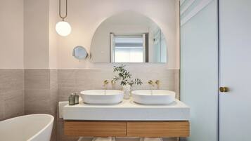 A modern bathroom with a freestanding bathtub, two sinks with gold faucets, a round mirror, and a light fixture.