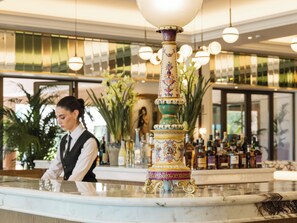 A hotel lobby with a marble reception desk, a decorative fountain, and a reflection of palm trees in the background.