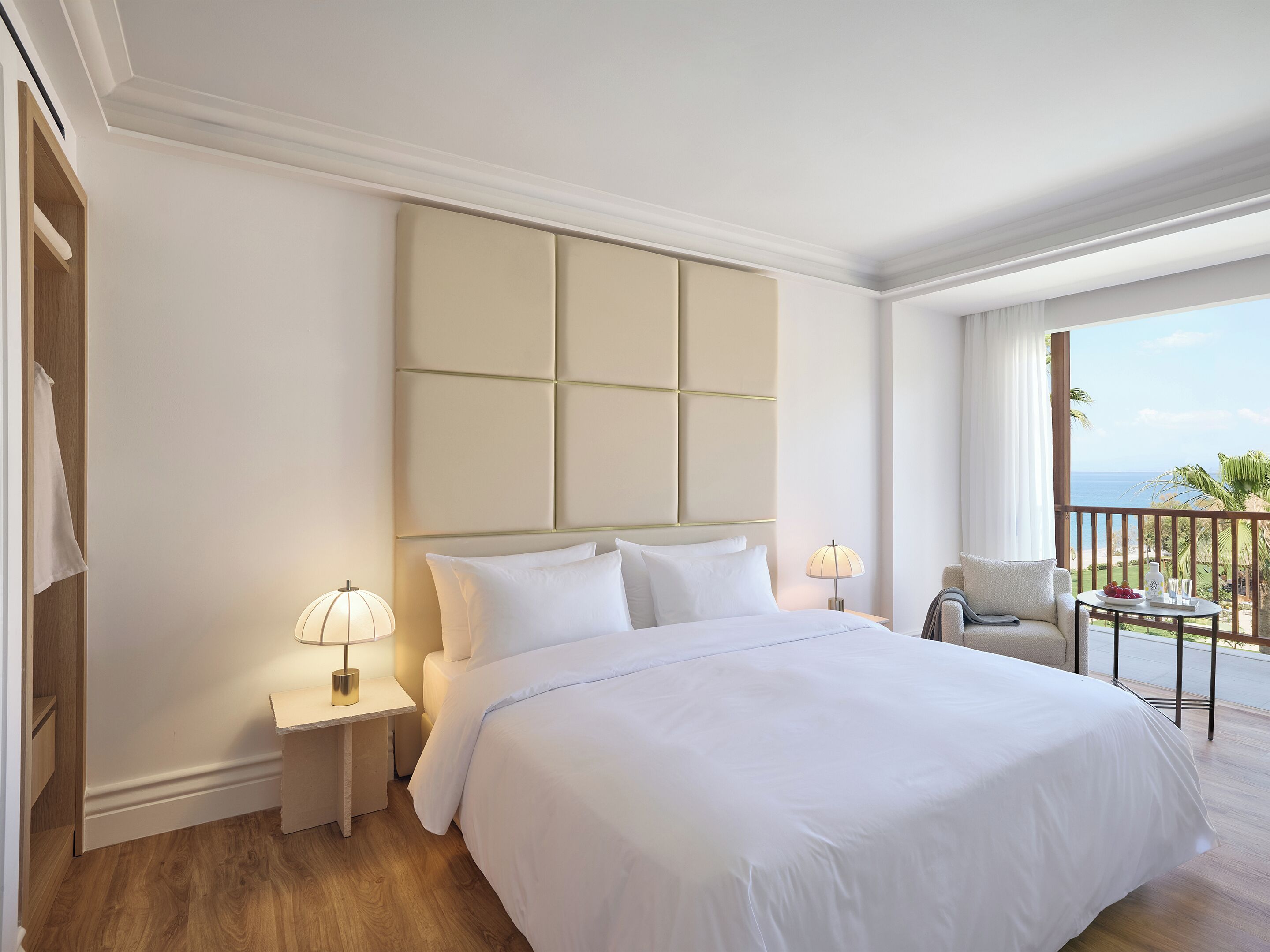A hotel room with a large bed, a nightstand with a lamp, a chair, and a small table with a view of the ocean.