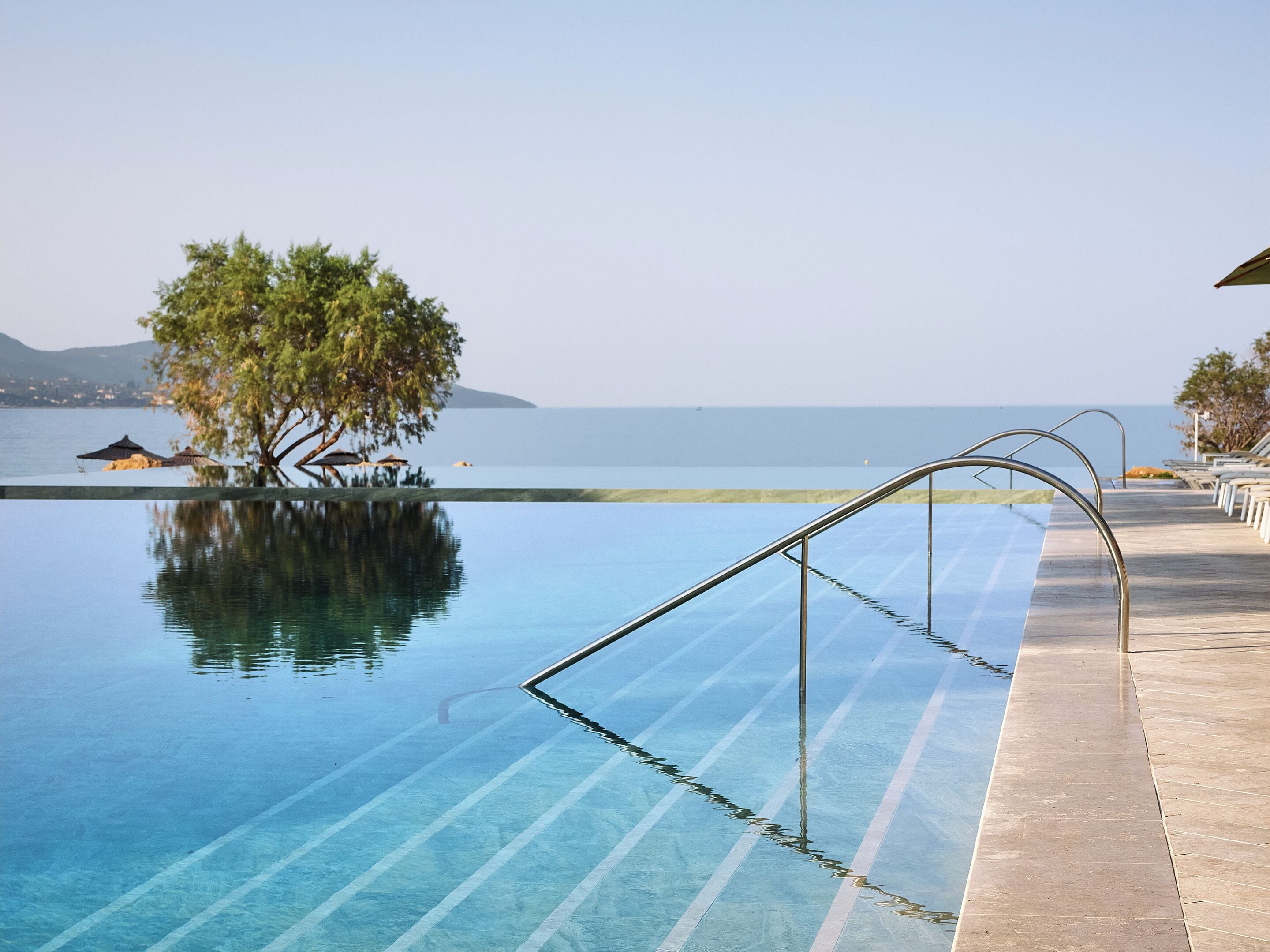 Ein klarer Infinity-Pool mit Holzterrasse und Liegestühlen, mit Blick auf ruhiges Gewässer und entfernte Berge.