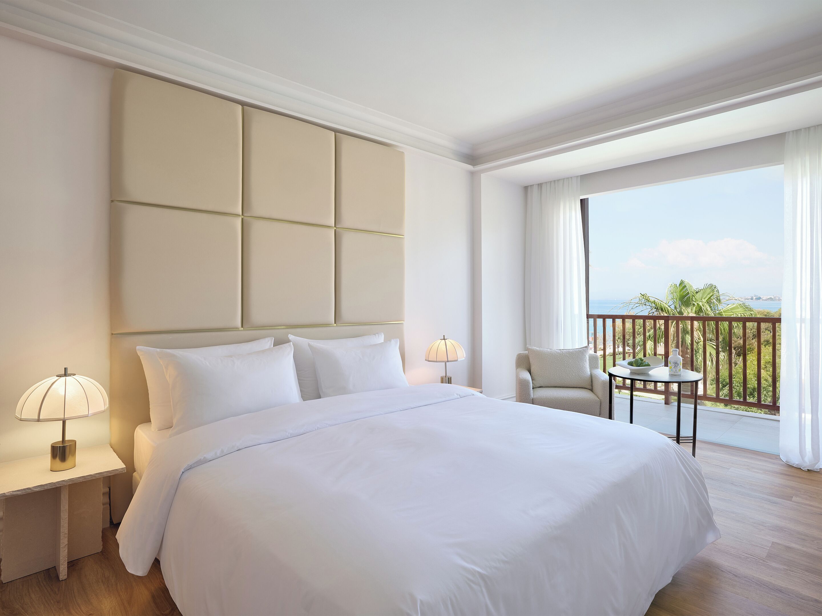 A hotel room with a large bed, a nightstand with a lamp, a small table with a chair, and a balcony with a view of the ocean.