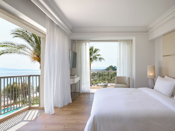 A hotel room with a large bed, a TV, a balcony with a view of the sea, and a small table with a lamp.
