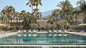 Zona de piscina con sillas de descanso, palmeras y un edificio con techo de tejas.