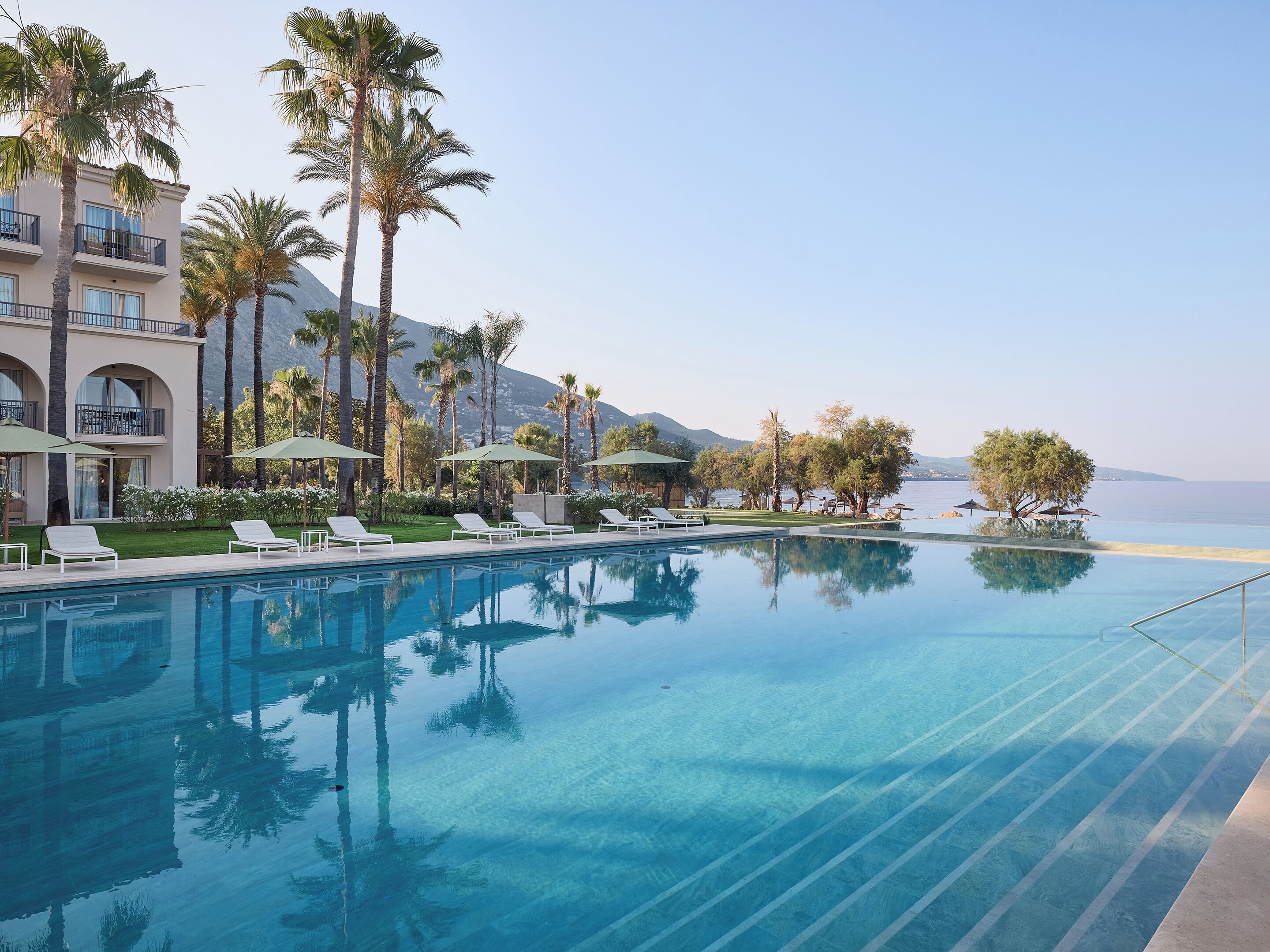 A pool with lounge chairs, palm trees, and a view of the sea.