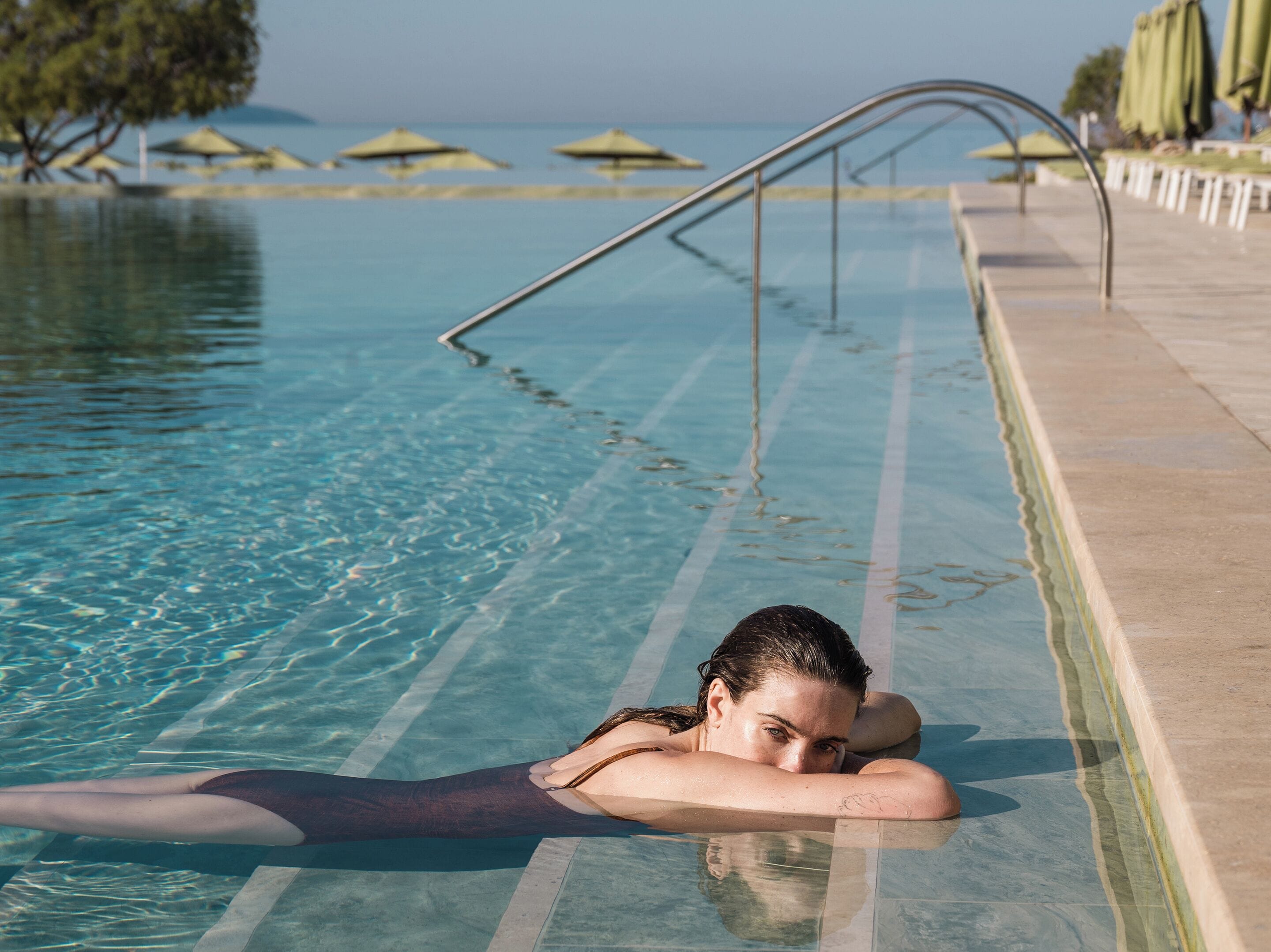 A pool with clear water, a poolside area with umbrellas, and a view of the sea.