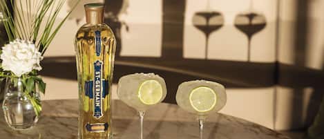 Una botella de St. Germain sobre una superficie de mármol, dos vasos con rodajas de lima y un florero con flores blancas.